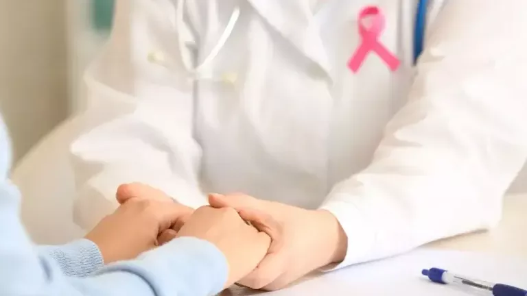 A healthcare professional wearing a white coat and a pink breast cancer awareness ribbon holds a patient’s hands in a supportive gesture during a consultation.