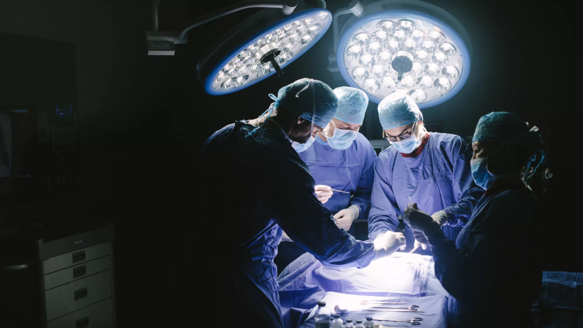 Surgical team in blue scrubs and protective masks performing a procedure in an operating theatre under bright surgical lights.